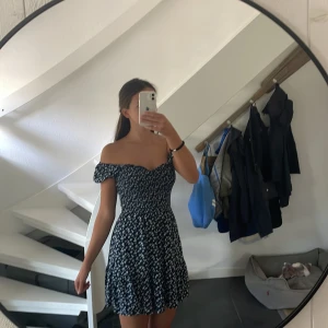 Blommig kortklänning från Hollister i storlek S - Supersöt marinblå kortklänning från Hollister med vita små blommor. Klänningen har smockad överdel, volangdetaljer och kan bäras både offshoulder och med puffärm. Mjuk och lätt bomull, perfekt för varma dagar. Säljer då den inte kommer till användning längre!