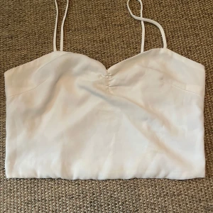 Vit siden bandeau topp med smala band - Supersnygg vit bandeau topp i siden med tunna axelband och lätt rynkning framtill. Toppen har en mjuk, glansig yta och en enkel, stilren look som är perfekt för varma dagar. Passar dig som gillar minimalistisk stil och vill ha något lätt och fräscht.