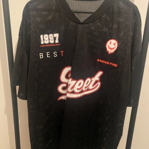 Svart mesh t-shirt från SHEIN XXL - Cool svart mesh t-shirt från SHEIN i storlek XXL. Oversized fit med tryck framtill: 'Street', '1997', och en röd smiley. Tillverkad i 100% polyester, perfekt för en sportig eller streetwear-look. Luftigt material och korta ärmar.