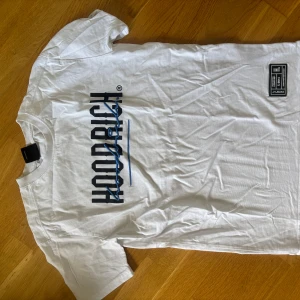 Vit HOODRICH t-shirt med blått tryck - Vit t-shirt från HOODRICH med stort blått och svart tryck både fram och bak. Klassisk rund hals och korta ärmar. Snygg streetwear-stil med coola detaljer och logga. Tillverkad i mjuk bomull för skön känsla hela dagen.