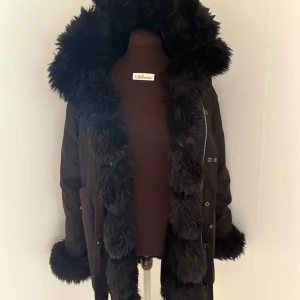 Svart parkajacka med fuskpäls - Snygg svart parkajacka med stor fluffig fuskpäls runt huva, ärmslut och längs öppningen. Jackan har dragkedja och knappar framtill samt praktiska fickor. Perfekt för kalla dagar när du vill vara både varm och stilren. VÄL ANVÄND