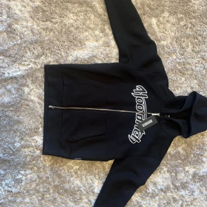 Svart hoodiejacka från Hoodrich  - Oanvänd Svart hoodiejacka från Hoodrich med vit broderad logga över bröstet. Jackan har dragkedja framtill, huva och två fickor. Tillverkad i mjukt bomullsmaterial, perfekt för en chill streetwear-look. 