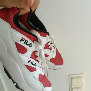 Rödvita sneakers från FILA - Snygga sneakers från FILA i rött och vitt med svarta detaljer. Skorna har en chunky sula, vit snörning och klassisk logga på sidan. Ovandelen är i textil och syntet med coola paneler och sportig vibe. Perfekta för dig som gillar streetstyle och vill sticka ut.