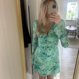 Grön Adoore klänning - Supersöt kortklänning från Adoore i gröna och turkosa toner med blommigt mönster. Klänningen har långärmade ärmar, v-ringad hals och figurnära midja. Perfekt för dig som vill ha en fräsch och färgglad look i sommar. 