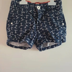 Marinblå shorts med ankare Hampton Republic - Snygga marinblå shorts från Hampton Republic med vitt ankarmönster och stjärnor. Shortsen har knappar fram och bak, uppvikta benslut och är tillverkade i 100% bomull. Perfekta för en somrig look med nautisk känsla.