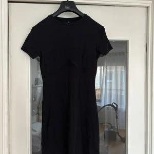 Svart ribbad kortklänning med söm - Svart ribbad kortklänning från Gina med rund hals och korta ärmar. Snygg detaljsöm under bysten som ger en cool form. Stretchigt material som sitter skönt och figurnära. Perfekt för dig som gillar minimalistisk stil med en twist.