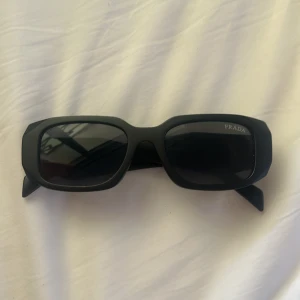 Svarta solglasögon från Prada - Säljer ett par svarta solglasögon från Prada Milano med breda skalmar och rektangulära bågar. Snygg och modern design med tydlig logga på sidan. Perfekta för dig som vill sticka ut med en lyxig accessoar. Kan förhandla om priset