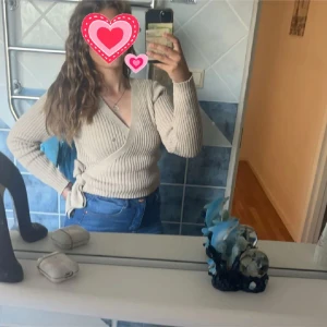 Beige omlott kofta med knyt - Säljer en beige ribbad omlottkofta med knyt i sidan. Tröjan har långärmade ärmar och v-ringad hals, perfekt att styla med jeans eller kjol. Materialet är mjukt och stickat, vilket gör den både skön och snygg. Passar dig som gillar en chill och trendig look.