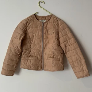 Tunn beige quiltad jacka från Bondelid strl.S - Quiltad beige jacka från Bondelid med rund halsringning och dragkedja framtill. Jackan har två fickor och diskret logga på bröstet. Perfekt för lager-på-lager och enkel att matcha med olika outfits.