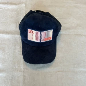 Blå keps All Cap One - Snygg mörkblå keps med mesh på sidorna och mockaliknande front. Framtill sitter en patch med ett 500-kronorssedel-motiv som ger kepsen en unik och edgy vibe. Justerbar baktill för perfekt passform. Kommer med svart påse med logga.