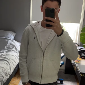 Ralph Lauren tröja - Säljer en stilren vit ziphoodie från Polo Ralph Lauren med en grön polo logga på bröstet. Den har luva då jag har dragit ut snörena. På sista bilden så ser man slitningar i armen som det är på båda ärmarna men som inte märks så länge men inte kollat nära. Perfekt för en chill outfit i sommaren och höst.🔥👑