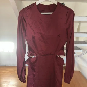 Vinröd omlottklänning Gina Tricot - Snygg vinröd klänning från Gina Tricot i omlottmodell med volangdetaljer och cut-outs vid midjan. Klänningen har lång ärm och rund halsringning, tillverkad i ett glansigt material som ger en lyxig känsla. Perfekt för dig som vill sticka ut med en unik look.