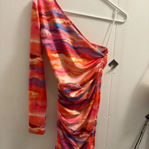 Färgstark oneshoulder kortklänning. Storlek M - Säljer en supercool kortklänning med oneshoulder och lång ärm. Klänningen har ett färgstarkt mönster i rosa, orange, lila och blå toner, med snygga rynkade detaljer längs sidan. Materialet känns mjukt och stretchigt, perfekt för att sticka ut.