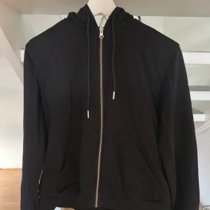 Svart hoodie med dragkedja från H&M - Svart hoodie från H&M i storlek M med hel dragkedja framtill, huva med snörning och två stora fickor. Tröjan har långa ärmar och är gjord i mjuk bomull, perfekt för chill dagar. Enkel och stilren design som funkar till det mesta.
