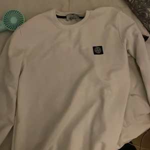 Vit Stone Island sweatshirt - Säljer en clean vit sweatshirt från Stone Island med klassisk patch på vänster ärm. Tröjan har rund hals och långa ärmar, perfekt för dig som gillar stilrena och exklusiva plagg. Materialet är mjukt och skönt, passar dig som vill ha en enkel men snygg look.passar s