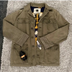 Olivgrön overshirt med stickad rygg - Snygg olivgrön overshirt med fyra stora fickor framtill och knäppning med knappar. Jackan har klassisk krage och raka ärmar. Perfekt för lager-på-lager och funkar till jeans eller chinos. Materialet är bomull och ger en avslappnad vibe.