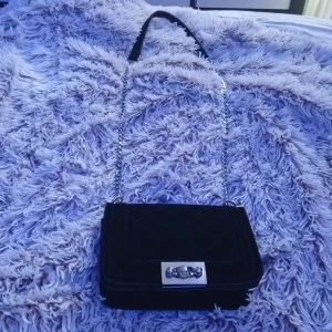 Svart quiltad axelväska Gina Tricot - Säljer en svart quiltad axelväska från Gina Tricot med kedjerem och snyggt silverfärgat spänne framtill. Väskan är i sammet och har ett innerfack med dragkedja. Perfekt storlek för det viktigaste och ger en stilren vibe till din outfit. Priset kan deskuteras 