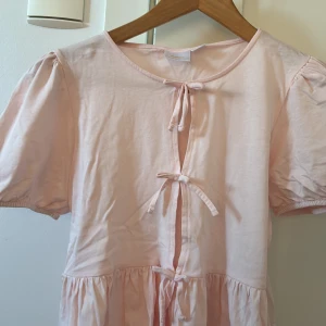 Ljusrosa topp från Gina Tricot - Supersöt ljusrosa topp från Gina Tricot med puffiga korta ärmar och tre rosetter framtill. Tillverkad i mjuk bomull och har en volang nedtill. Den är i storlek 158/164