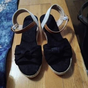 Svarta sandaler med rosett, storlek 36 - Säljer ett par snygga svarta sandaler med bred rosett framtill och plattformssula i repimitation. De har beige textilrem med brunt spänne runt ankeln och en vit räfflad undersida. Perfekta för dig som gillar en chill och trendig sommarstil. Helt ny aldrig använts men tyvärr har jag inte kvittot