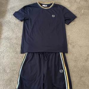 Sergio Tacchini tracksuit i storlek S - Mörkblå tracksuit från Sergio Tacchini med ljusblå och gula ränder längs sidorna. Shortsen har snörning i midjan och är tillverkade i ett lätt, ventilerande polyestermaterial. Perfekta för sommaren, med klassisk logga på benet.