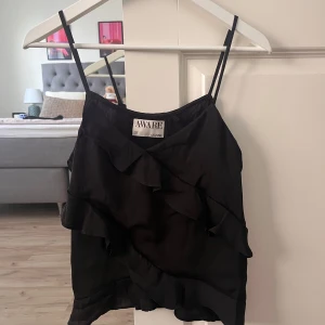 Svart linnetopp med volanger från AWARE - Svart linnetopp från AWARE med tunna axelband och snygga volangdetaljer framtill. Toppen har en luftig och lätt känsla, perfekt för varma dagar. Stilren design som ger en cool vibe och passar till många olika looks.