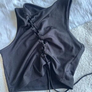 Svart ribbad snörad topp Divided S - Säljer en svart ribbad topp från Divided, H&M i storlek S. Toppen har snörning framtill som man själv kan välja hur hårt knytet det ska vara. Endast använd en gång