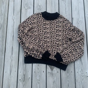 Leopardmönstrad beige tröja från Lindex  - Säljer en snygg leopardmönstrad tröja i beige och svart med svarta muddar vid hals, ärmslut och nederkant. Tröjan har långärmade, lite puffiga ärmar och en loose passform. 
