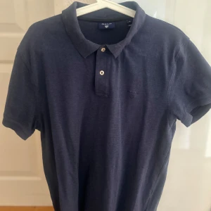 Mörkblå GANT pikétröja XL - Snygg mörkblå pikétröja från GANT i storlek XL. Klassisk krage med två knappar och diskret broderad logga på bröstet. Tillverkad i mjuk bomull som känns skön mot huden. Perfekt för dig som gillar stilrena och tidlösa plagg.