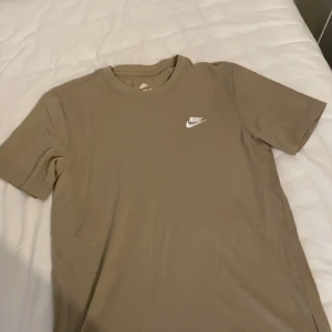 Beige Nike t-shirt i bomull - Snygg beige t-shirt från Nike med klassisk logga i vitt på bröstet. Modellen har rund hals och korta ärmar, tillverkad i mjuk bomull som är skön mot huden. Perfekt för dig som gillar enkel och stilren streetwear.