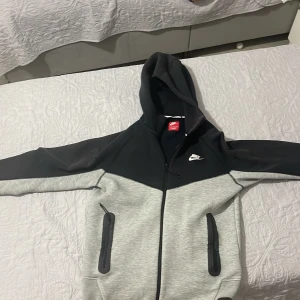 Nike hoodiejacka svart/grå S - Nike hoodiejacka i svart och ljusgrått med dragkedja framtill och två fickor med dragkedja. Jackan har huva och klassisk Nike-logga på bröstet. Perfekt för chill eller träning, tillverkad i mjuk bomullsblandning.