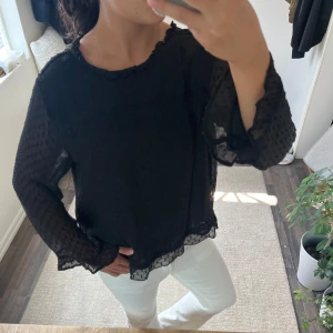 Svart prickig blus med volangkant - Säljer en svart blus med diskret prickigt mönster och transparenta, luftiga långärmar. Blusen har volangkant vid halsringning och nederkant, vilket ger en extra snygg touch. Perfekt att styla med jeans eller kjol för en cool look.