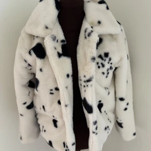 Vit och svart pälsjacka med dalmatinermönster - Säljer en fluffig pälsjacka i vit med svarta dalmatinerliknande fläckar. Jackan har bred krage och stängs med tryckknappar. Perfekt för dig som vill sticka ut och hålla dig varm med stil. 