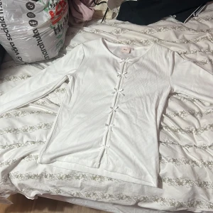 Vit ribbad långärmad topp från Nelly - Supersöt vit ribbad topp från Nelly i storlek S. Långärmad med flera små rosetter framtill som ger en gullig vibe. Materialet är mjukt och stretchigt, perfekt för dig som gillar bekväma och stilrena plagg. Passar dig som vill ha en enkel men ändå detaljrik look.