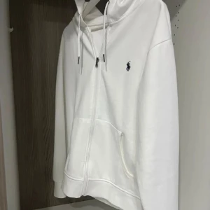 Vit hoodie från Polo Ralph Lauren - Snygg vit hoodie från Polo Ralph Lauren med dragkedja och huva. Klassisk broderad logga på bröstet och praktiska fickor framtill. Tillverkad i mjuk bomull, perfekt för en clean och stilren look. Passar dig som gillar enkel men exklusiv streetstyle.