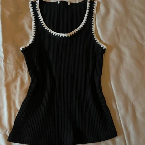 Svart ribbad linnetopp med vit spetskant - Snygg svart ribbad linnetopp med vit broderad kant runt halsringning och ärmhål. Perfekt för en stilren look.