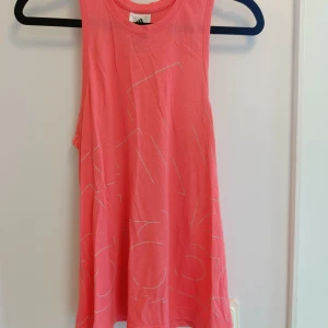 Rosa linnetopp från Adidas - Snygg rosa linnetopp från Adidas med vita grafiska linjer. Toppen är ärmlös och har en avslappnad passform, perfekt för träning eller varma dagar. Mjukt bomullsmaterial som känns skönt mot huden.