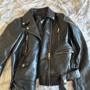 Svart skinnjacka med bälte  - Snygg svart skinnjacka med klassisk bikerstil, flera dragkedjor, bred krage och bälte i midjan. Jackan har coola detaljer som tryckknappar och fickor med dragkedja. Perfekt för dig som vill ha en edgy look.