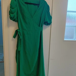 Grön omlottklänning med puffärm - Snygg grön långklänning med omlottmodell och puffiga kortärmar. Klänningen har v-ringning och knytband i midjan för en smickrande siluett. Perfekt för dig som gillar färg och vill sticka ut lite extra.