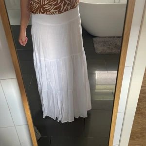 Vit långkjol från Chica London i storlek S - Supersnygg vit långkjol från Chica London i luftig bomull. Kjolen har bred smock i midjan som går att vika ner och flera volanger som ger en flowy och chill vibe. Perfekt för varma dagar och enkel att matcha med crop tops eller linnen.