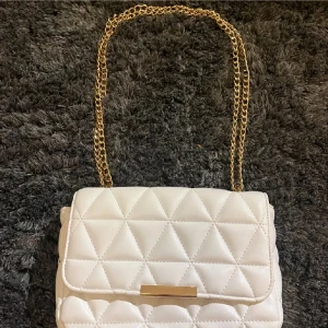 Vit quiltad axelväska med guldkedja - Snygg vit quiltad axelväska med elegant guldkedja. Väskan har en praktisk dragkedja inuti och en stilren design. Perfekt för att ge din outfit en lyxig touch.