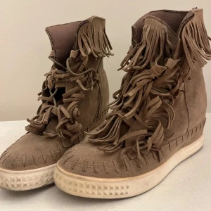 Bruna mocka boots med fransar - Snygga bruna boots i mocka med fransdetaljer som ger en bohemisk touch. Skorna har snörning framtill och en bekväm sula med mönstrad kant. Perfekta för en avslappnad stil.