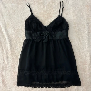 Svart spetstopp från La Senza - Säljer en elegant svart topp från La Senza med spetsdetaljer och en söt rosett framtill. Toppen har justerbara axelband och en vacker spetskant längst ner. Perfekt för en speciell kväll!