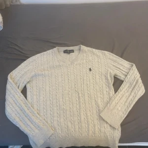 Beige stickad tröja från Ralph Lauren - Säljer en stilren beige stickad tröja från Ralph Lauren med klassiskt flätmönster. Tröjan har en v-ringad hals och långa ärmar, perfekt för kyligare dagar. Den ikoniska loggan är broderad på bröstet. Passar både till vardag och mer uppklädda tillfällen.