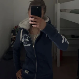 Blå hoodie från Superdry - Säljer en snygg blå hoodie från Superdry med vit text på ärmen och dragkedja framtill. Den har en mysig fodrad huva och är perfekt för kyliga dagar. Passar både till jeans och mjukisbyxor. Perfekt för en avslappnad stil! 💙