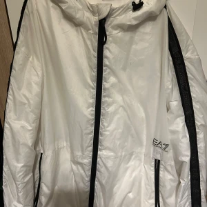 Vit vindjacka från Emporio Armani - Säljer en stilren vit vindjacka från Emporio Armani med svart dragkedja och huva. Jackan har en sportig look med EA7-logga på bröstet och svarta detaljer längs ärmarna. Perfekt för vår och sommar. Passar även för dem som har M. Pris kan diskuteras.
