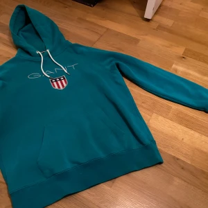 Turkos hoodie från GANT - Säljer en snygg turkos hoodie från GANT med vit logga och emblem på bröstet. Tröjan har en klassisk känguruficka och justerbar huva med vita snören. Perfekt för en avslappnad stil.