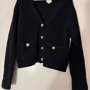 Svart ribbad kofta med knappar - Snygg svart ribbad kofta med guldknappar framtill och två fickor. Perfekt för att hålla sig varm och stilren. Passar bra till både jeans och kjol.