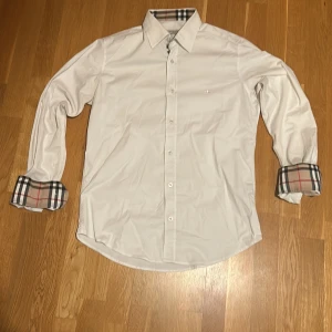 Vit skjorta från Burberry - Snygg vit skjorta från Burberry med klassiskt rutigt mönster på insidan av kragen och ärmsluten. Skjortan har långa ärmar och knappar framtill. Perfekt för en stilren look.💯 Den är bara testad och har inga defekter eller tecken på användning alls✅ Om du har några frågor eller annat är det bara att höra av dig😊 (Priset är inte hugget i sten men inga skambud)