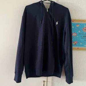 Svart hoodie från Ralph Lauren - Säljer en stilren svart hoodie från Ralph Lauren med dragkedja och huva. Tröjan har långa ärmar och en liten vit logotyp på bröstet. Perfekt för en avslappnad look.
