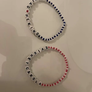 Två pärlarmband med text - Två handgjorda pärlarmband med bokstavspärlor. Det ena armbandet har blå och vita pärlor med texten 'JAG <3 PEKING'. Det andra armbandet har rosa och vita pärlor med texten 'EVIGHET CAROLA'. Perfekta för att uttrycka personlighet och stil.